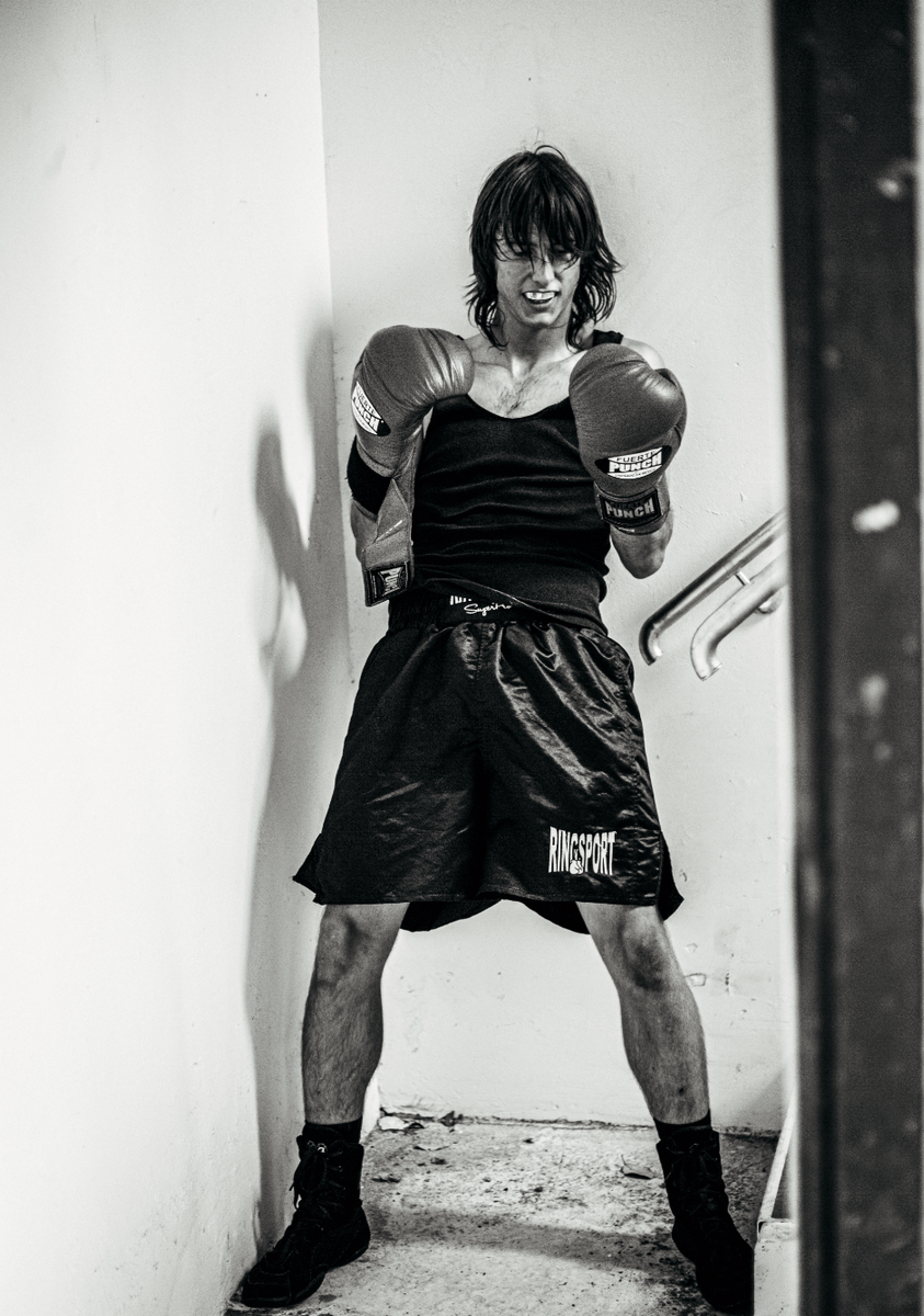 Black and white portrait photograph of a person in boxing stance wearing branded boxing gloves and shorts in a narrow white concrete corridor.