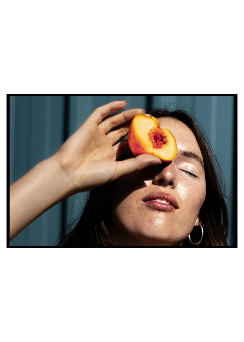Portrait photograph of a woman holding a halved peach in front of her face, with bright natural light creating warm skin tones against a soft teal-blue background.